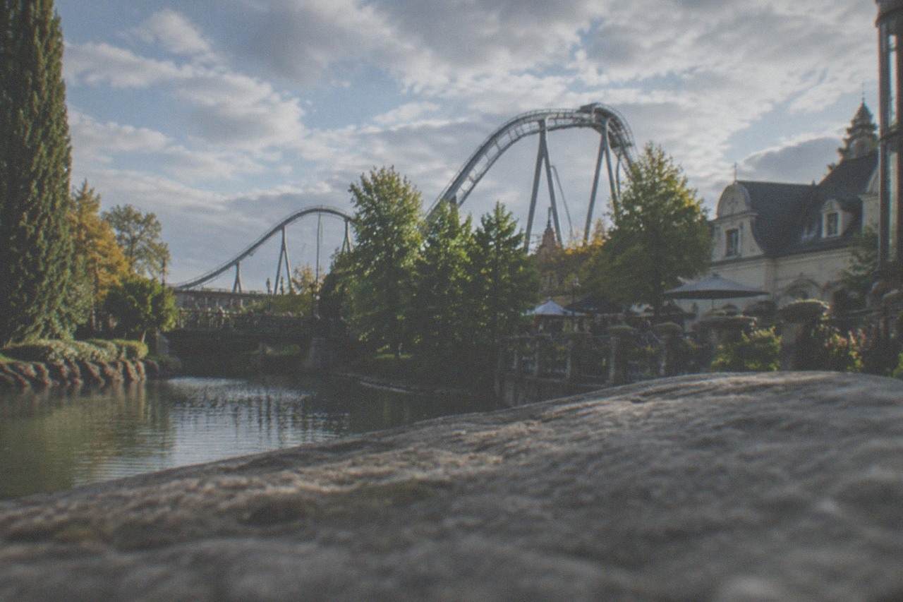 Roller coaster track rising above the treetops of a large European theme park under a blue sky