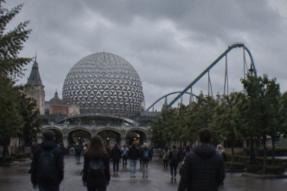 Theme park entrance plaza with a large silver sphere building and visitors walking toward the turnstiles
