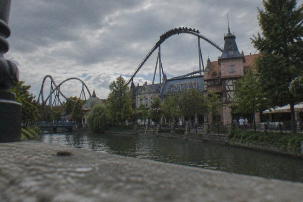 Roller coaster track curving over the themed rooftops of a large amusement park under a summer sky