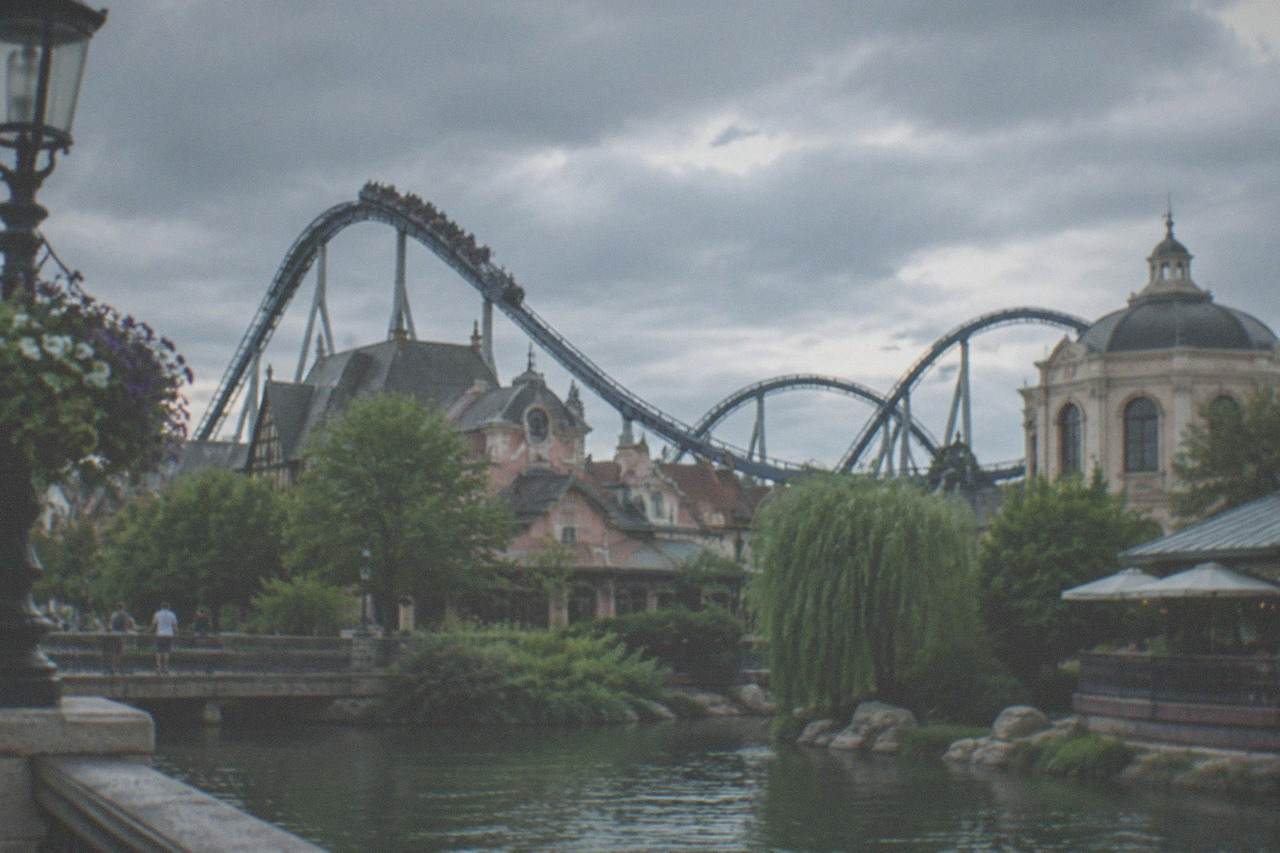 Roller coaster track curving over the themed rooftops of a large amusement park under a summer sky