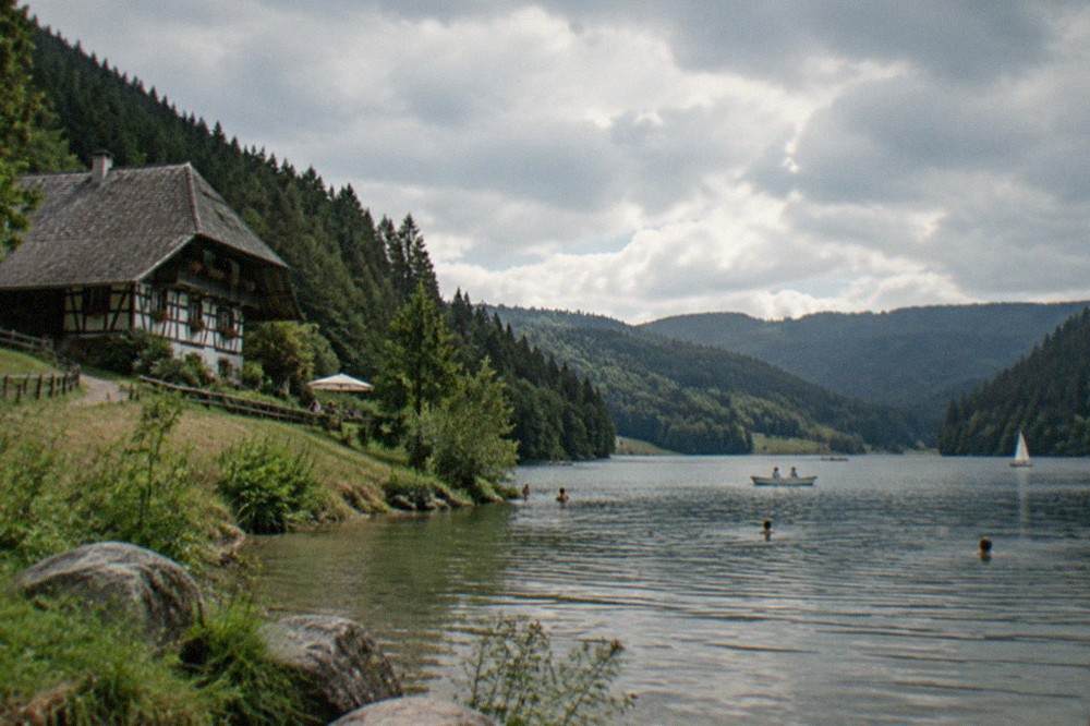 Swimmers and small boats on a calm lake surrounded by dark forest hills in summer