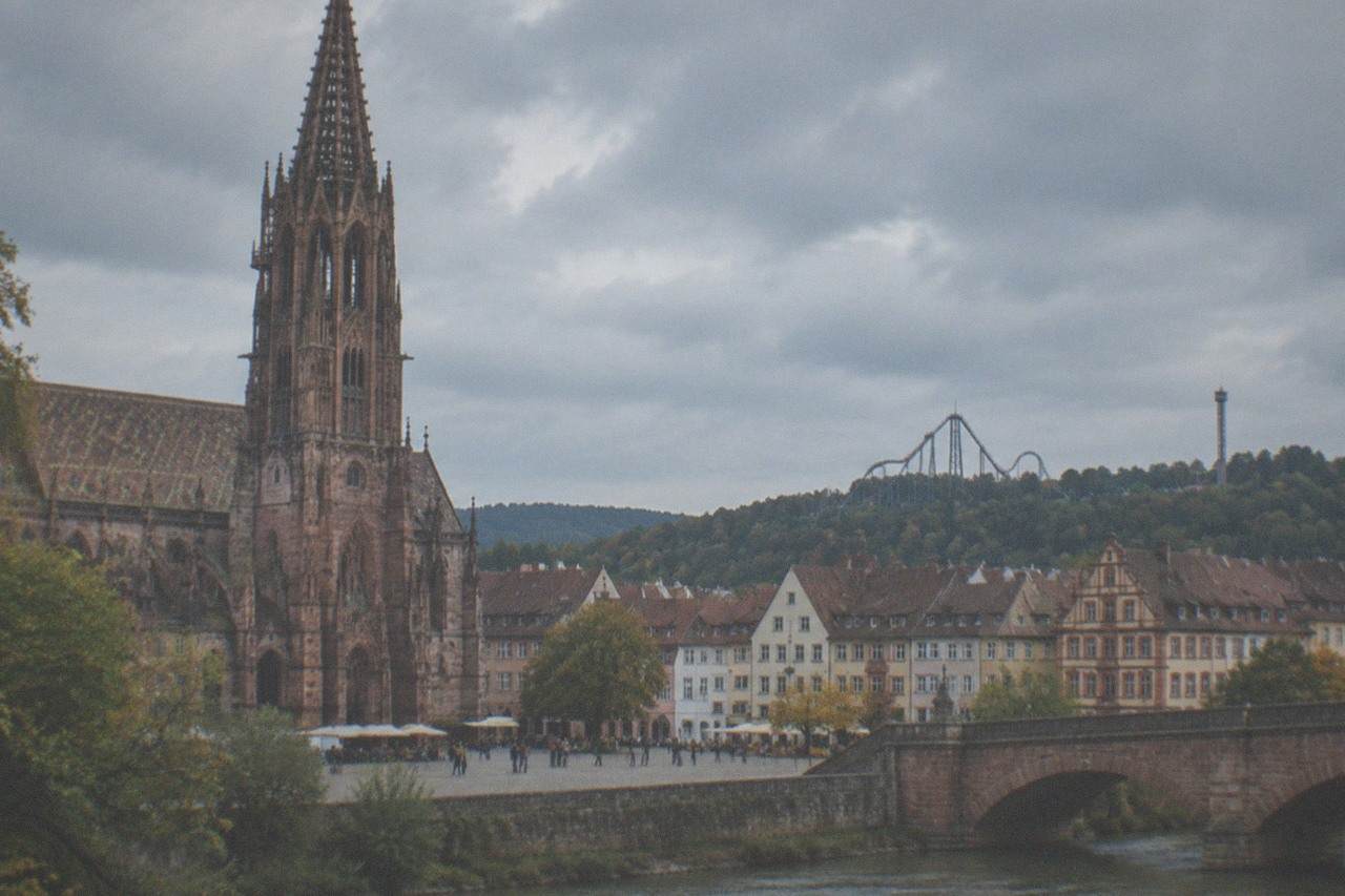 Roller coaster track rising over a European theme park skyline with forested hills behind