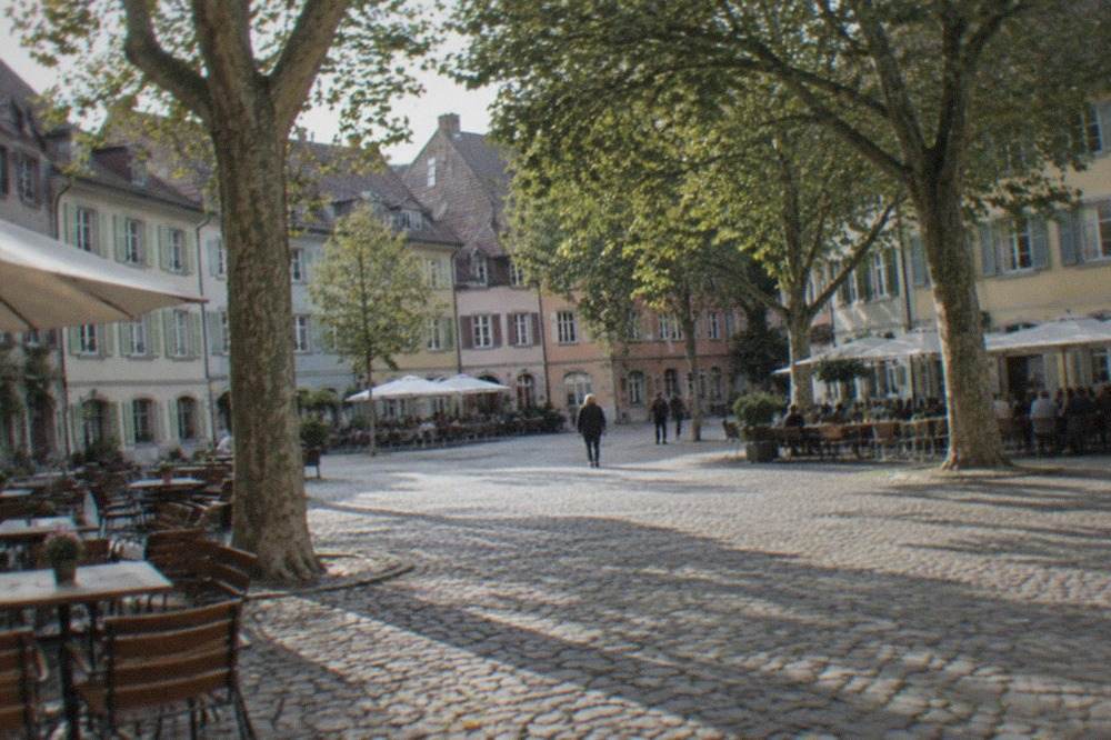 Wide cobblestone square with plane trees, outdoor tables and old town facades