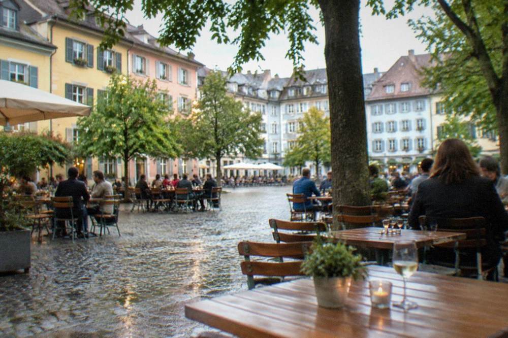 Evening terrace with drinkers and diners at wooden tables under plane trees