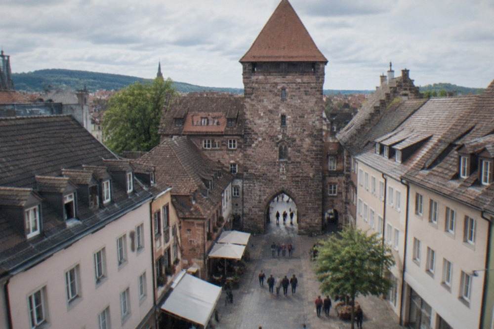 A tall stone gate tower with a steep red roof above a pedestrian street with shoppers