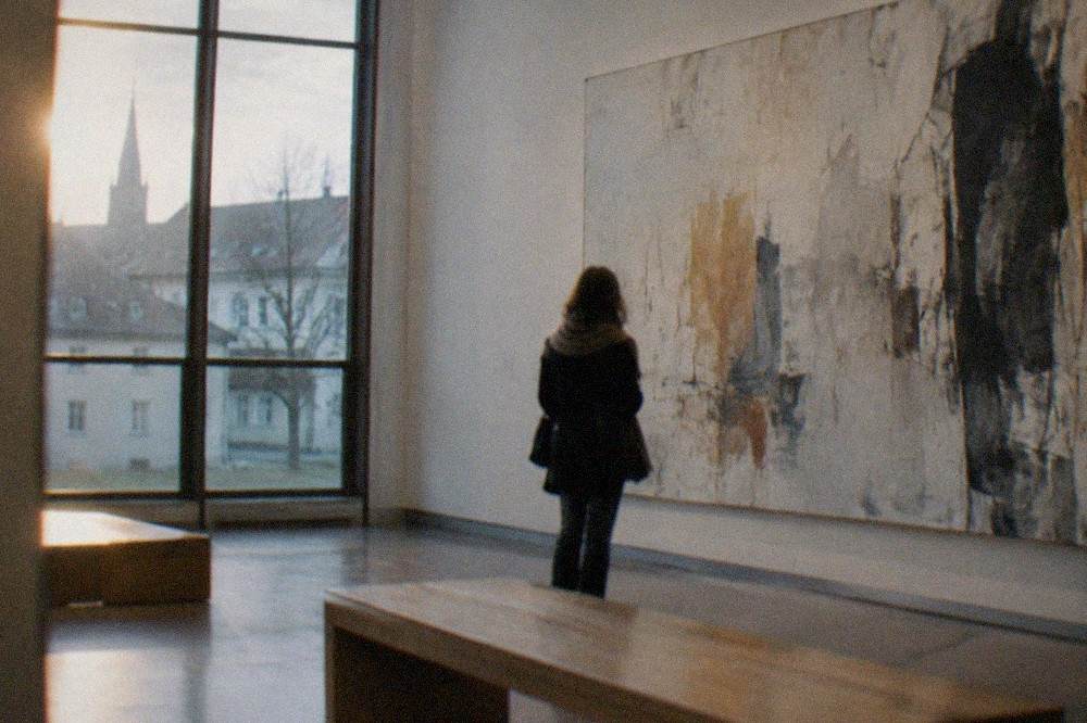 Visitor viewing a large abstract canvas in a quiet contemporary art gallery with wooden benches.