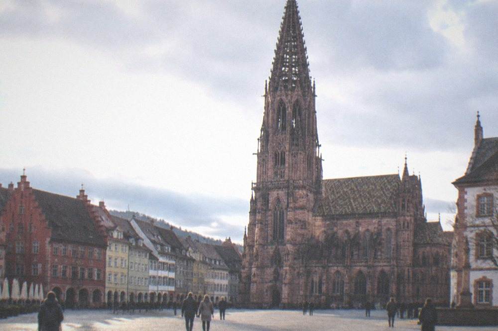 Wide view of a Freiburg square with historic guild facades and a gothic tower under a clear sky.