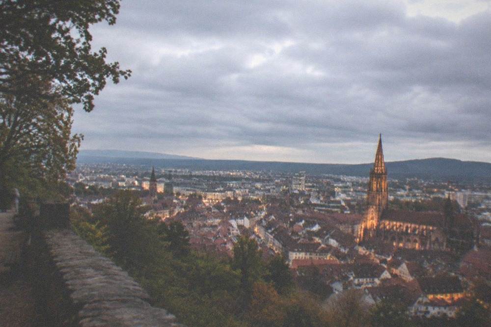 Evening cityscape seen from a hilltop terrace with lit rooftops and a floodlit cathedral tower