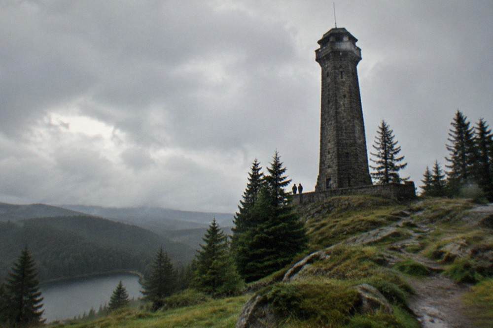 A stone viewing tower standing above fir forest on a high open ridge under scattered clouds.