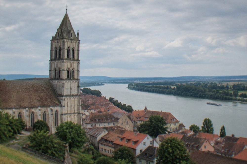 View over a small old town with a large stone minster tower rising above red rooftops toward a wide river plain