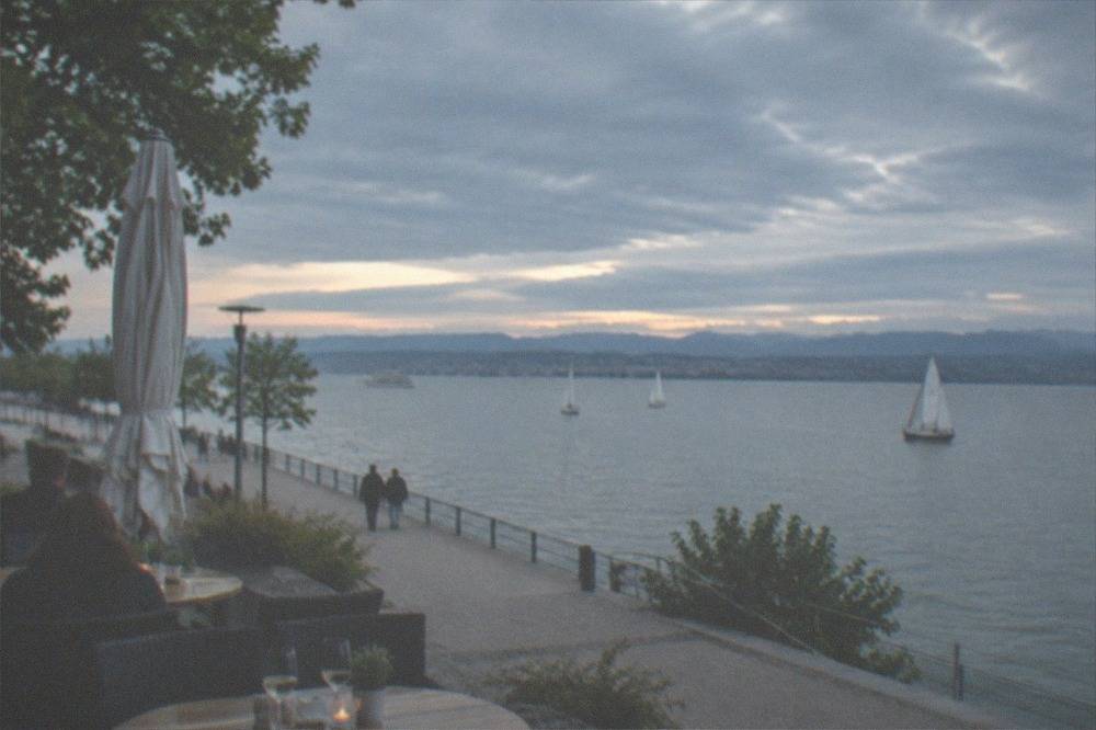 Outdoor cafe tables beside a lake promenade with water and boats in the background