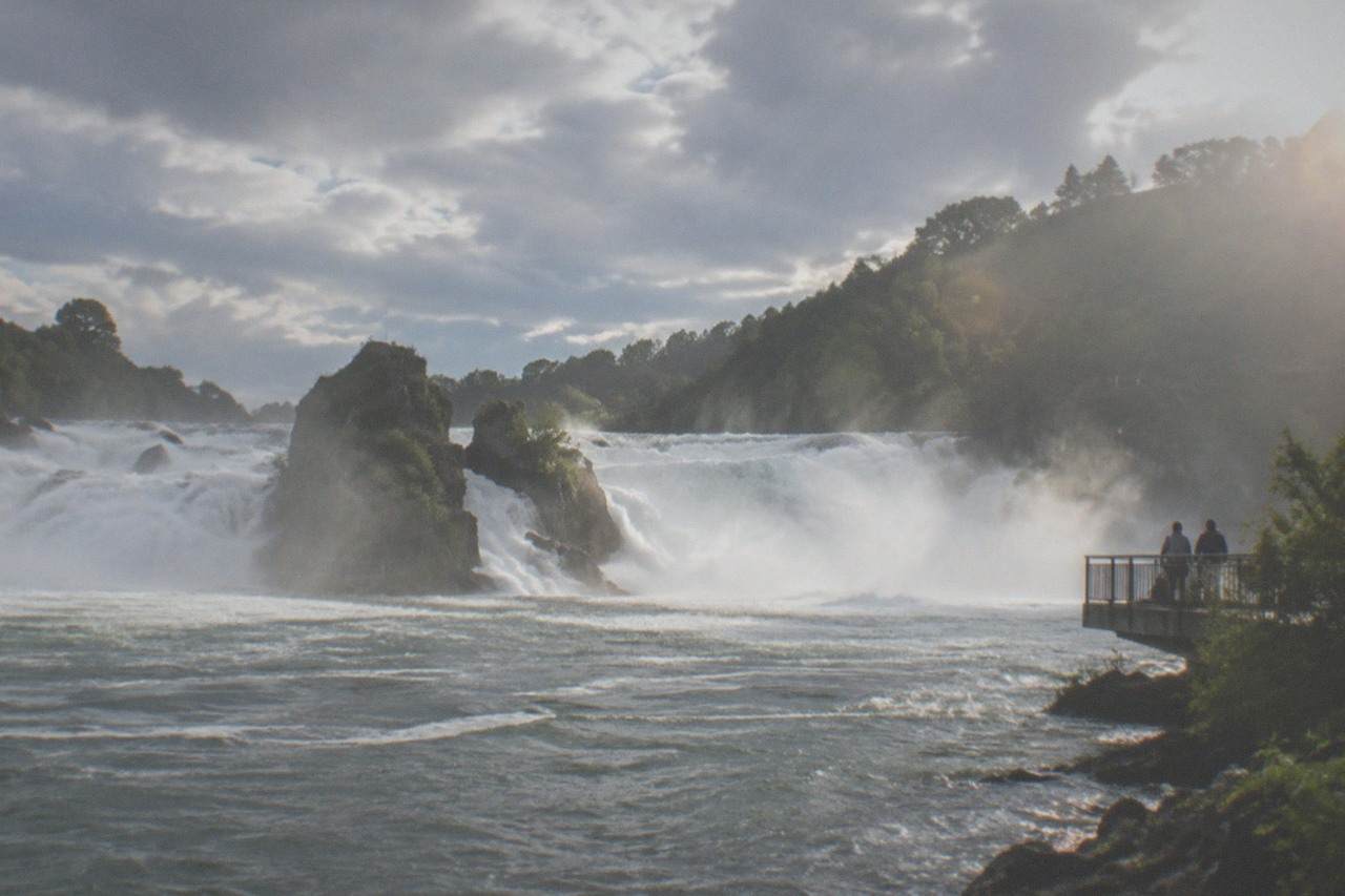 Wide rocky gorge with a large waterfall pouring between forested banks under a summer sky