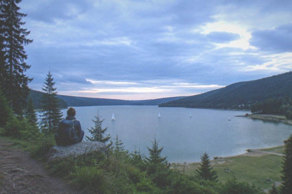 A hiker with a backpack resting on a forest path above a wide lake