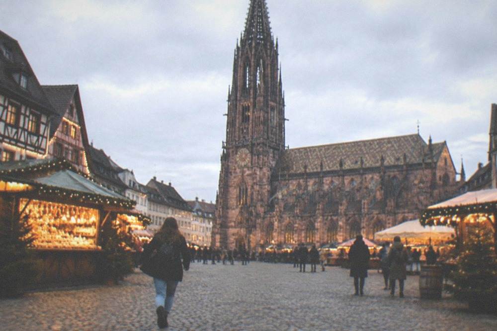 A festively lit old town square in Freiburg at dusk with market stalls and timbered facades