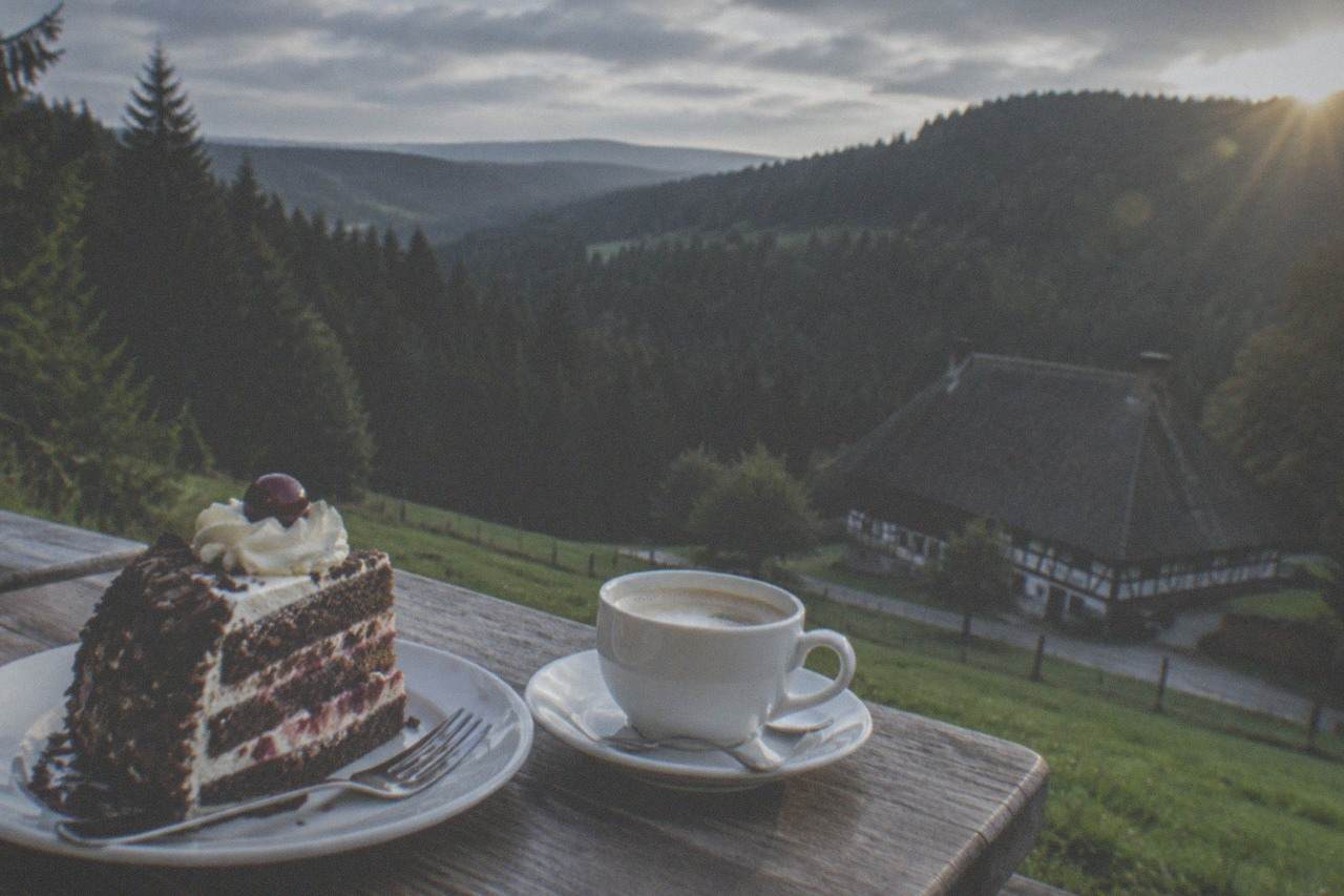A slice of Black Forest cake with cream, cherries and chocolate shavings on a café plate beside a coffee cup