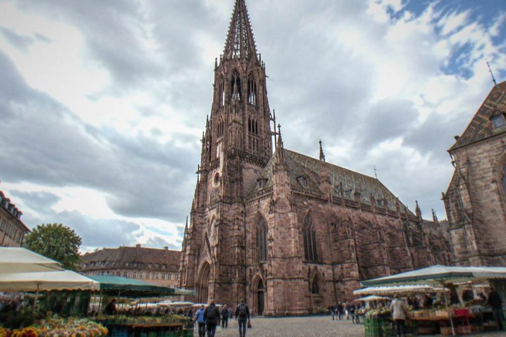 Market stalls under umbrellas beside a Gothic cathedral tower on a bright morning