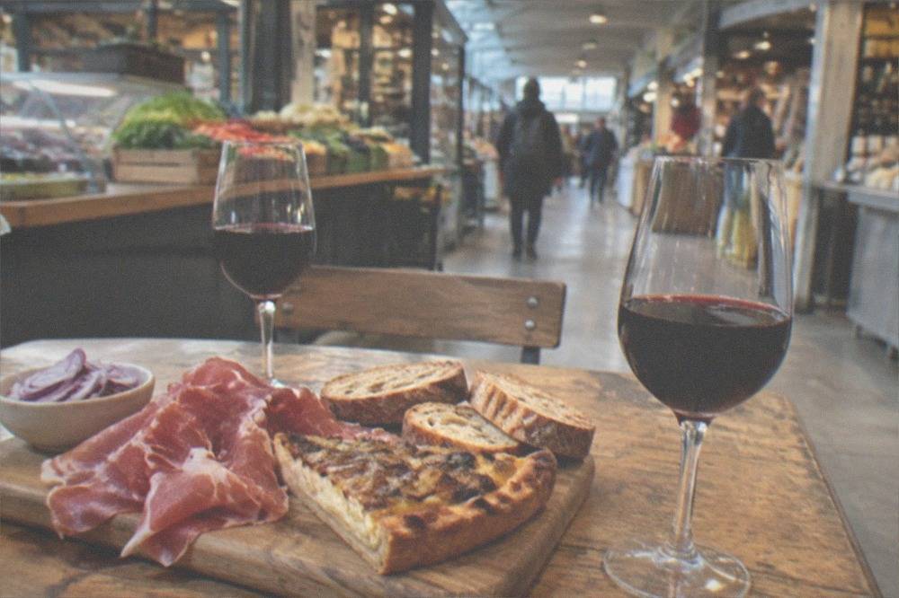 A tavern table set with a wooden board of ham, onion tart, bread and two glasses of red wine