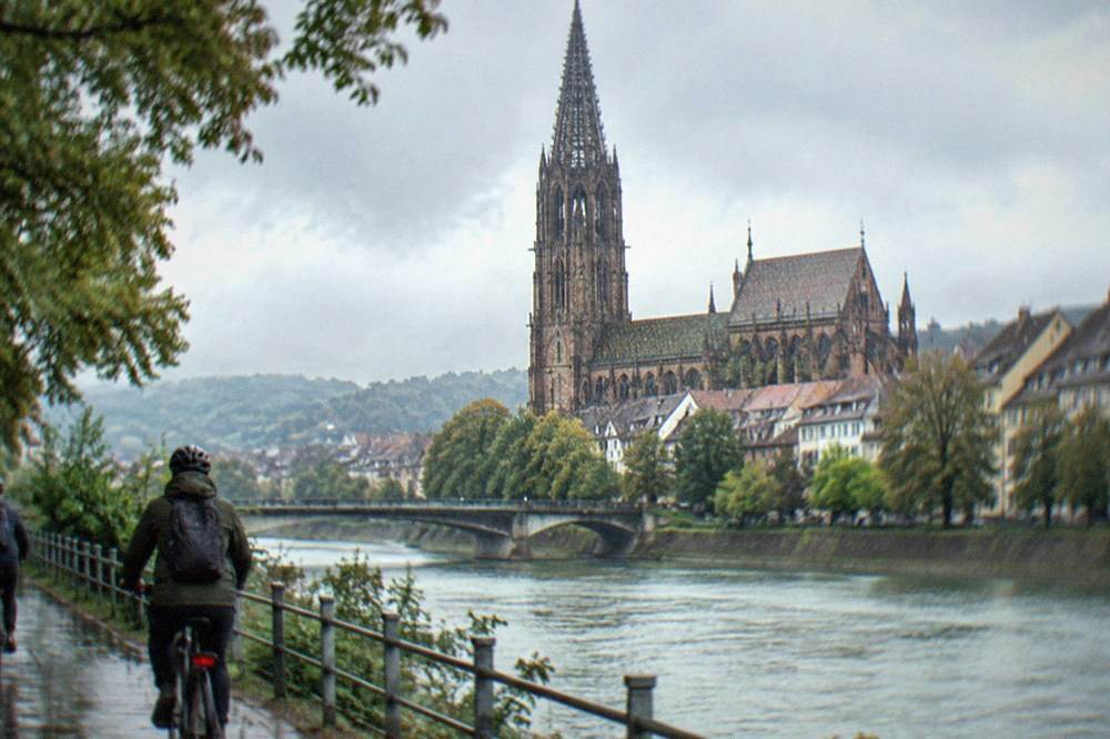 Cyclists on a riverside path with the Freiburg Minster visible in the background