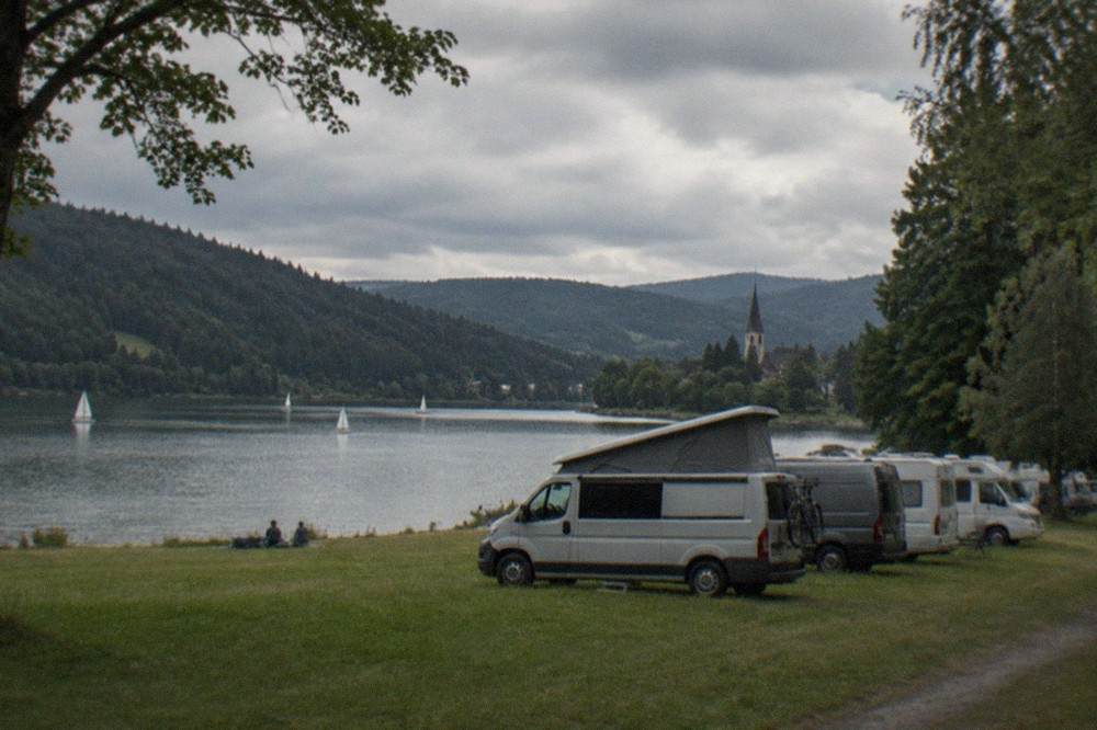 Campervans parked in a row on a tree-lined field with a church spire in the distance