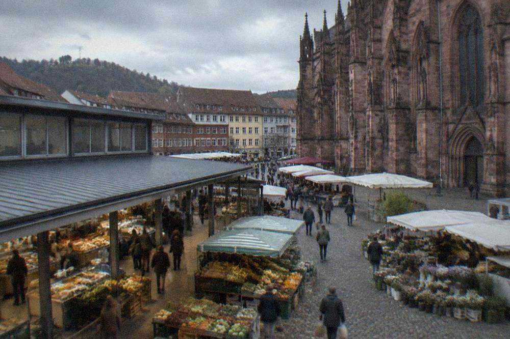 Produce stalls with vegetables, bread and flowers spread across a square beside a red sandstone cathedral