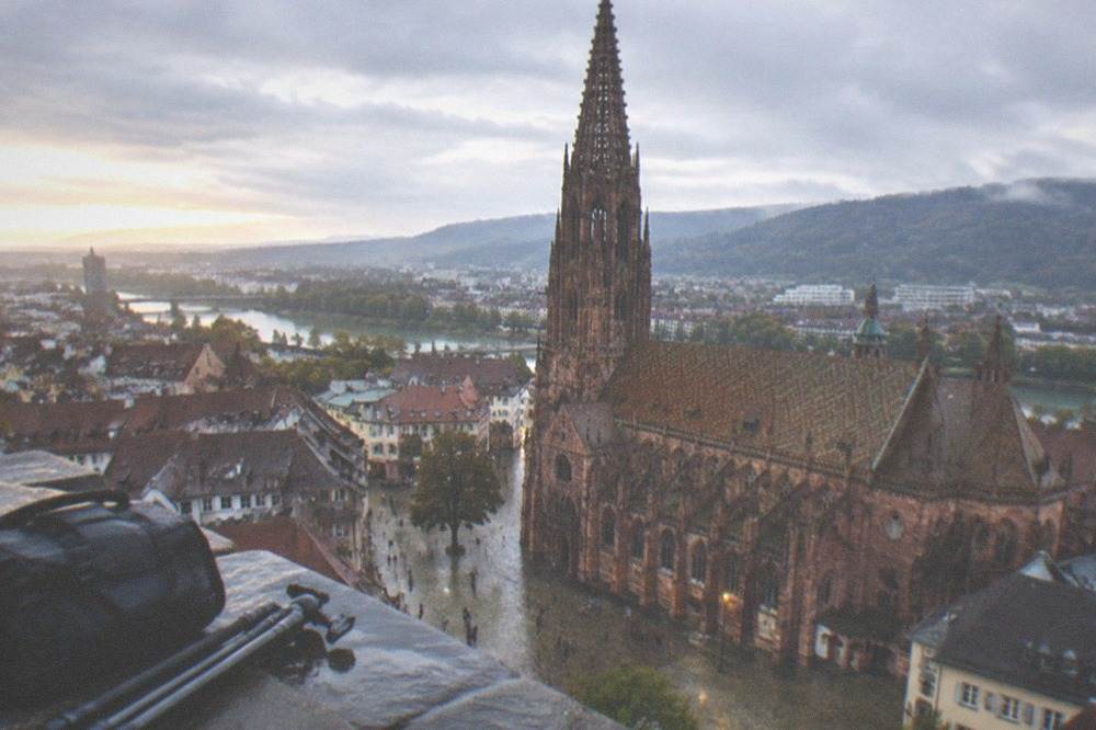 A photographer's camera bag and tripod on a stone wall overlooking Freiburg rooftops at dusk
