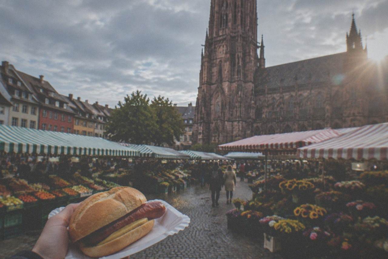 Münstermarkt stalls in front of Freiburg Cathedral with a red sausage in a bread roll on a paper plate.